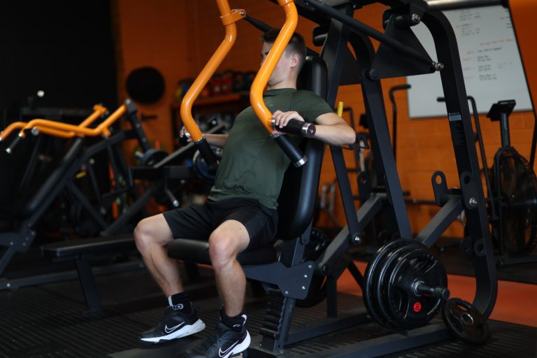 Man exercising on a seated shoulder press machine in a gym with orange and black decor.