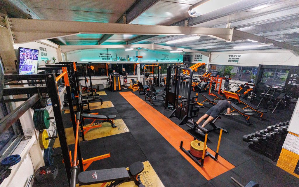 A gym features various workout equipment, including weight machines, benches, and free weights. Two individuals are exercising; one is using a leg press machine, and another is on a squat rack. A television is mounted on the wall showing a sports game.