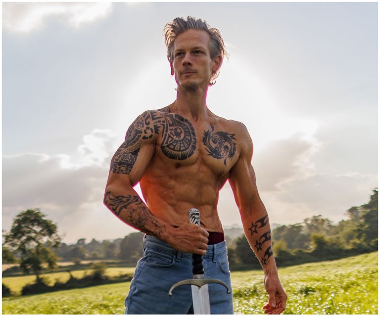 A shirtless man with tattoos, wearing jeans, stands in an open field holding a sword. The sun is shining behind him, casting a glow. Trees are visible in the background, and the sky is partly cloudy.