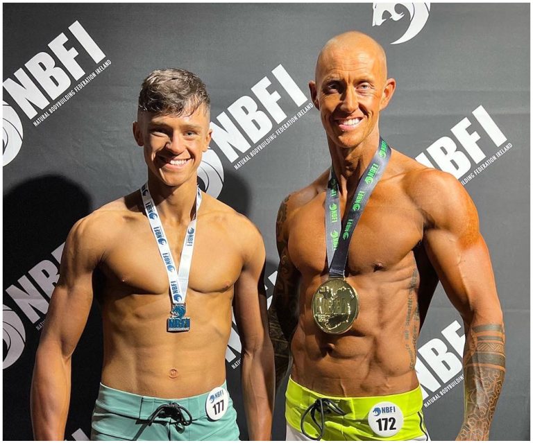 Two men stand side by side with medals around their necks in front of an NBFI backdrop. They are shirtless and smiling at the camera, with one wearing teal shorts (left) and the other wearing neon yellow shorts (right). The man on the right has a medal labeled "172".