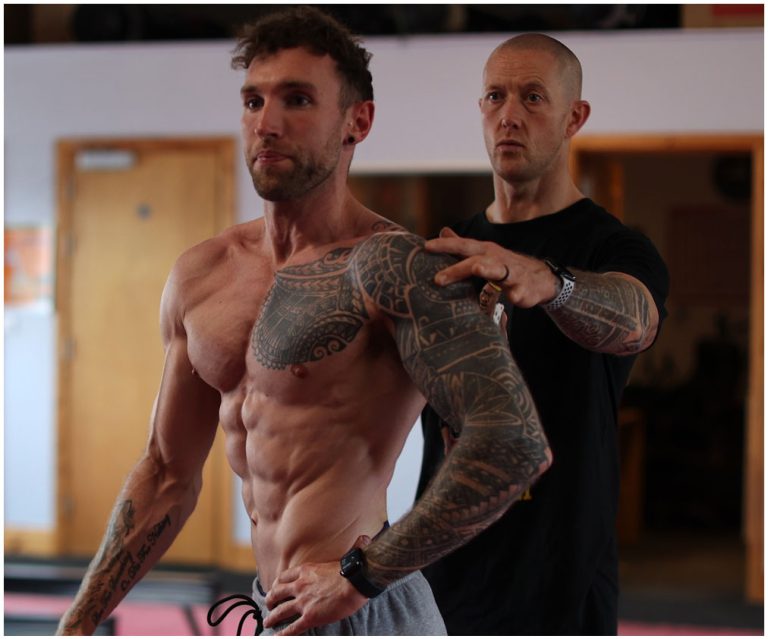 Two men are in a gym. One is shirtless, showcasing a muscular physique with extensive tattoos, and the other, standing behind him in a black t-shirt, appears to be a trainer, guiding or instructing him with one hand on his shoulder and the other on his back.