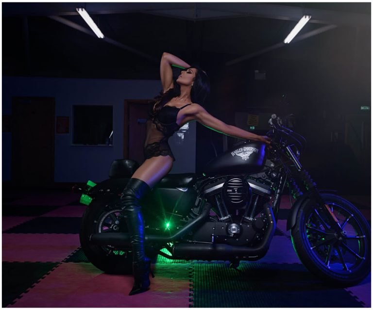 A woman in a black lingerie outfit and thigh-high boots poses with her head tilted back, standing beside a black Harley-Davidson motorcycle in a dimly lit room with overhead lights and colorful floor tiles.